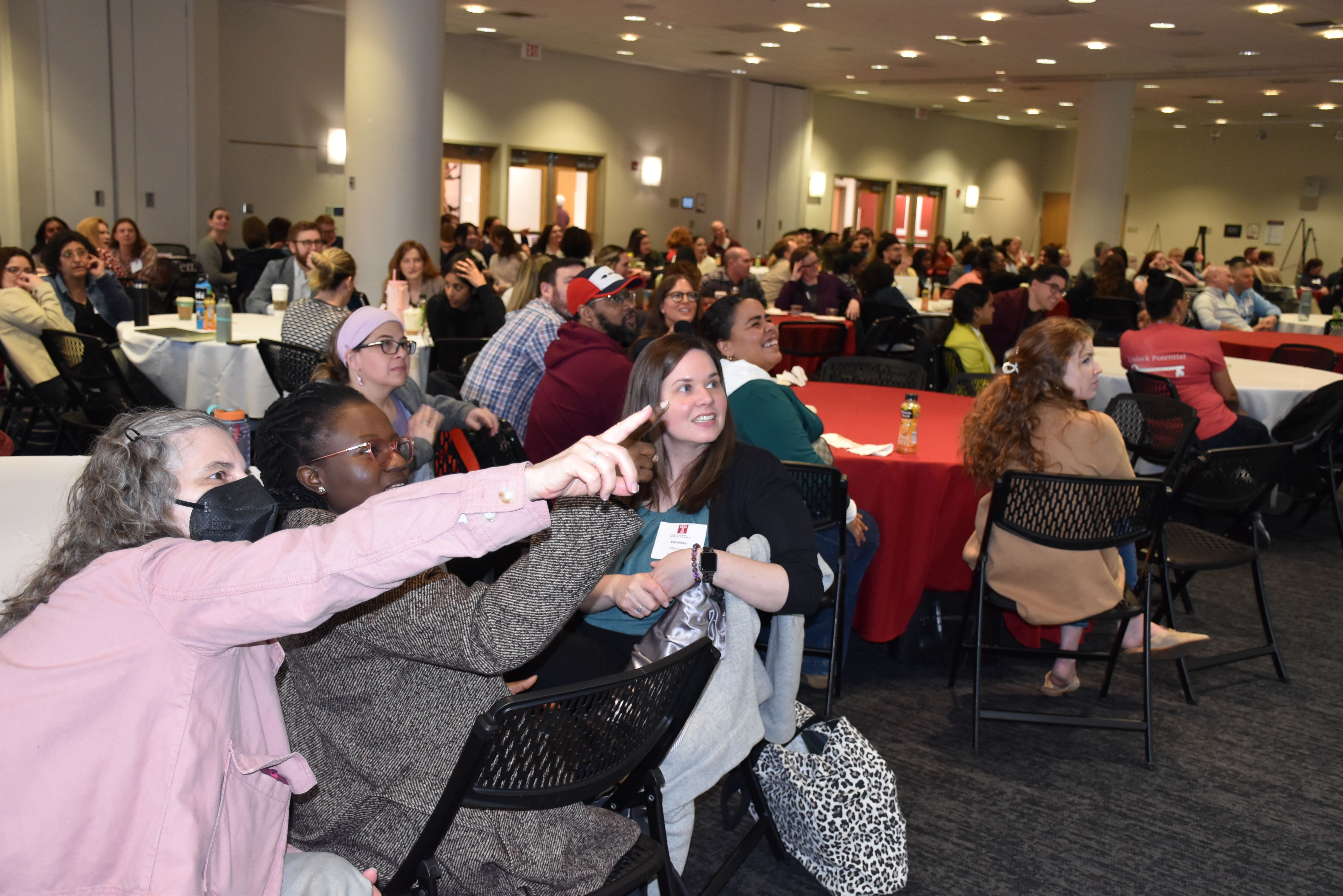 Wide shot of conference participants during a session. They are sitting at round tables and one person in the foreground has her arm outstretched, pointing toward what is being presented. 