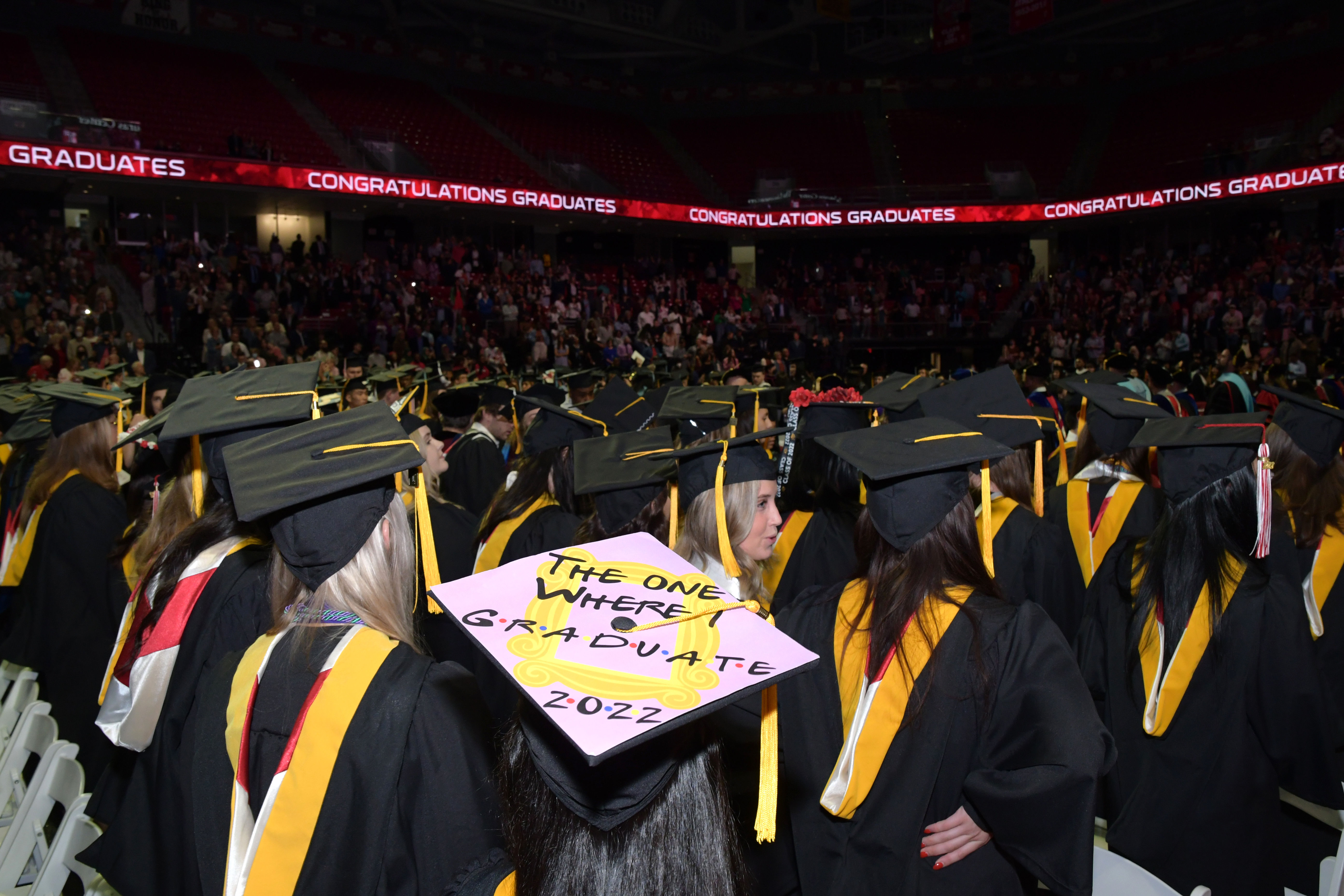 graduates with backs to camera in arena with "congratulations graduates" electronic signage