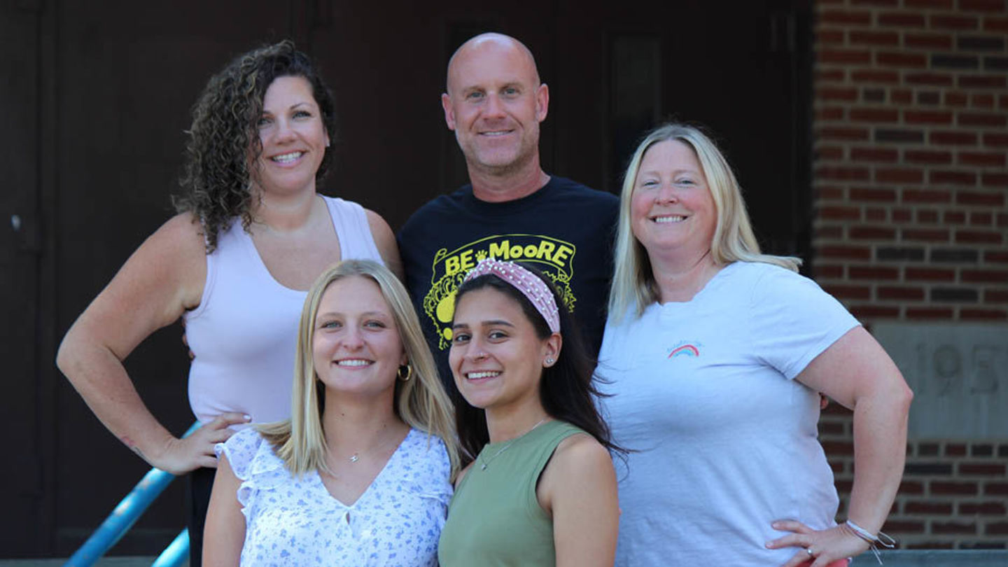 Pictured left to right: First row, Temple University 2022 graduates Jaclynn Campos and Hailey Lucas; second row, J. Hampton Moore Elementary School Assistant Principal Candice Lord (EDU '08), Principal Tim Glynn, School-Based Teacher Leader Kim Speed. Five people standing on the steps outside a brick building with brown doors. Three in the back are school administrators. Two in the front are recent Temple graduates.