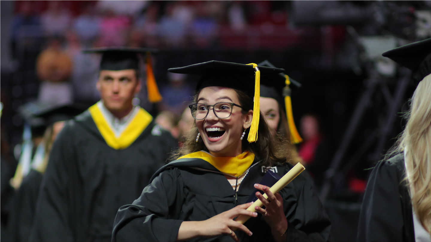 Graduating student in cap and gown with a wide, open-mouthed smile, holding a rolled up paper like a diploma