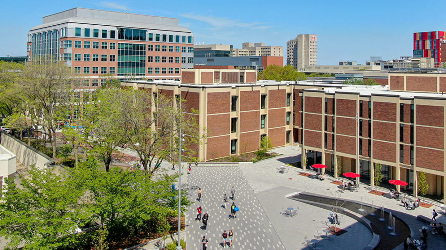 Aerial view of students walking on brick-paved path near Ritter Hall, a brick building. The sky is bright blue. There are trees by the path and a water feature.