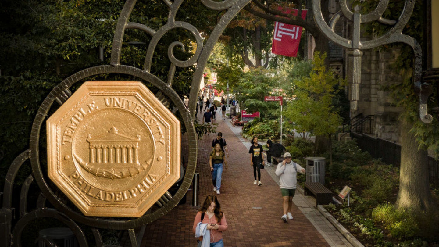 Aerial view of the gateway entrance to Temple University showing wrought-iron gate with bronze seal, and looking down at people walking on a tree-lined brick path with a red Temple T flag peeking out from the side.