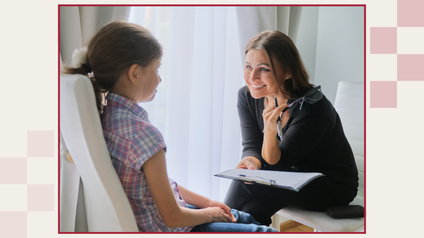 A woman is seated holding a clipboard while leaning in to speak with a school-aged child, also seated.ed child.