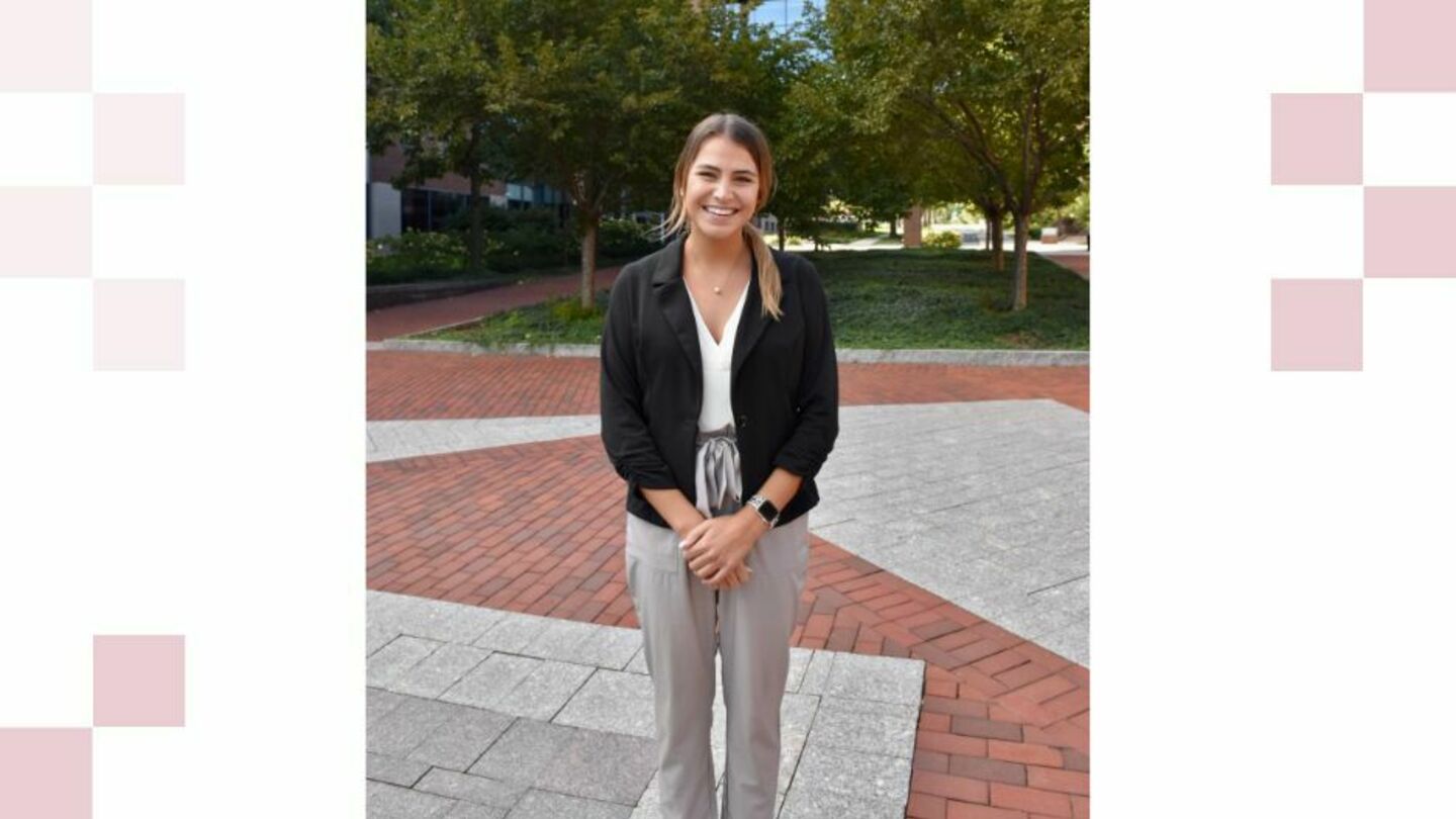 Emily Duddy stands wearing professional attire on a brick walkway with lush green trees in the background