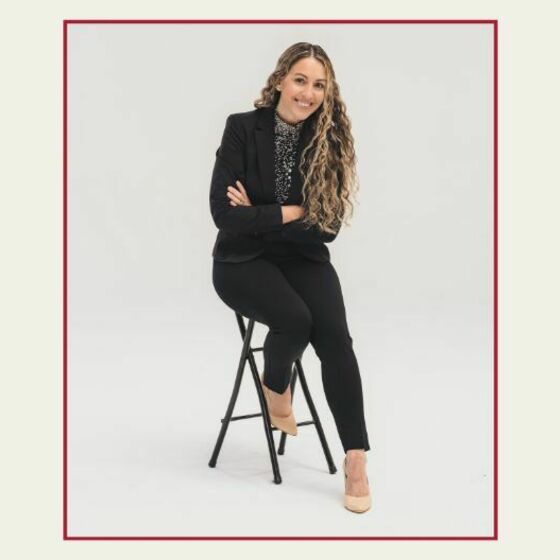 Amberly Caballero in a dark suit poses leaning forward on a high chair