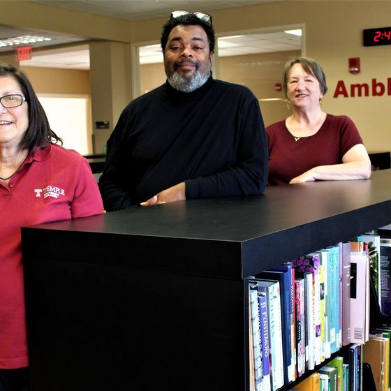 Three members of Ambler Campus Library Staff stand beside a bookcase.
