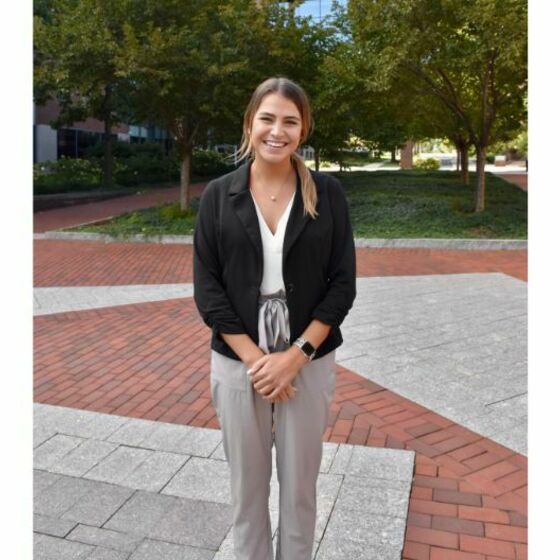 Emily Duddy stands wearing professional attire on a brick walkway with lush green trees in the background